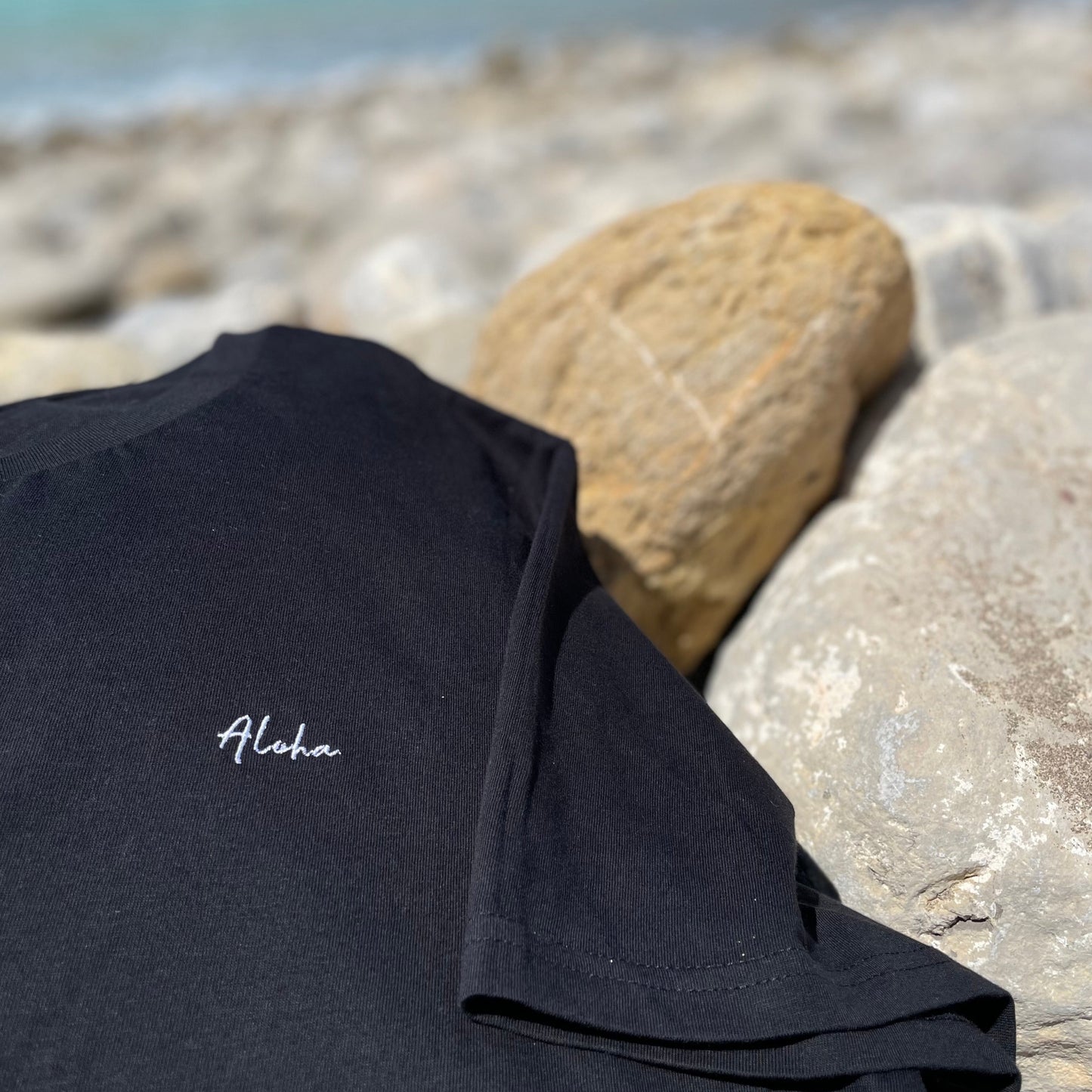 Nachhaltige Strandmode: Schwarzes 'The Aloha' T-Shirt auf Steinstrand